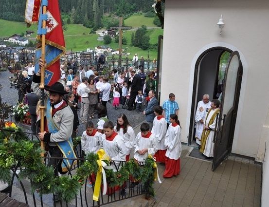 Eine Gruppe von Menschen, darunter Kinder in weißen Gewändern, versammelt sich vor einer Kirche mit einem Fahnenträger und einem Priester.