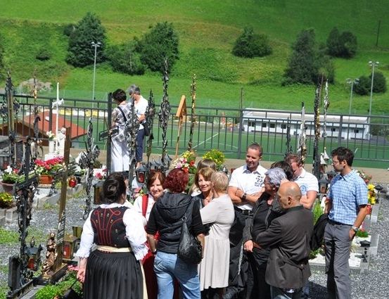 Eine Gruppe von Menschen auf einem Friedhof, einige in traditioneller Kleidung, stehen um ein Grab mit Blumen und einer Statue. Im Hintergrund sind ein grünes Feld und ein Gebäude zu sehen.
