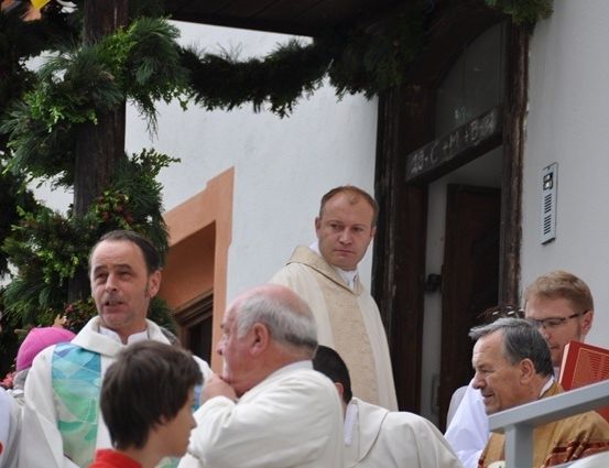 Eine Gruppe von Männern in weißen Roben, möglicherweise Priester, steht vor einem Gebäude. Ein Mann hält das Ohr eines Jungen.