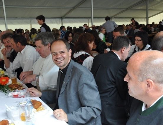 Eine Gruppe von Menschen sitzt an einem langen Tisch in einem Zelt und genießt eine Mahlzeit. Einige lächeln und andere führen Gespräche. Der Tisch ist mit Tellern, Gläsern und Blumen gedeckt.