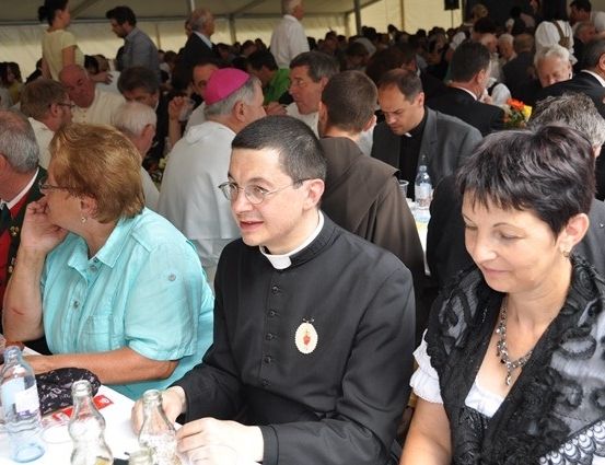 Ein Priester in einem schwarzen Gewand mit einem weißen Kragen sitzt an einem Tisch mit einer Frau. Sie sind von anderen Menschen in einer großen Versammlung umgeben, möglicherweise bei einer Kirchenveranstaltung.