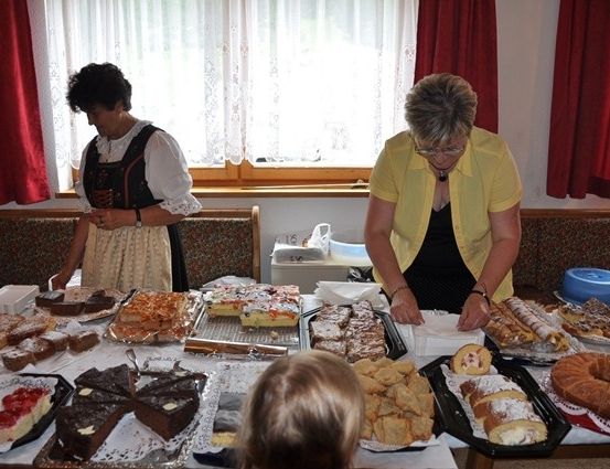 Zwei Frauen in traditioneller und lässiger Kleidung bereiten eine Vielzahl von Desserts auf einem Tisch zu, während ein Kind im Vordergrund beobachtet. Die Umgebung umfasst ein Fenster mit roten Vorhängen und einer weißen Tischdecke.