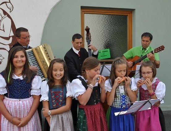 Eine Gruppe junger Mädchen in traditionellen Kostümen spielt Flöten, während sie vor einem Gebäude mit einem Wandbild stehen. Dahinter spielt ein Mann Akkordeon, ein anderer Cello und ein Dritter Gitarre.