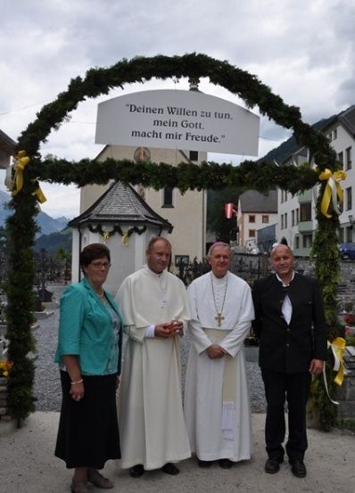 Vier Personen stehen unter einem dekorativen Torbogen mit einer deutschen Inschrift vor einem Kirchengebäude.