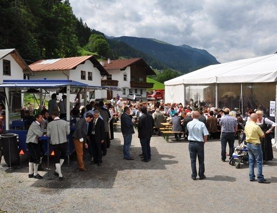 Eine Gruppe von Menschen versammelt sich auf einem Dorfplatz, einige stehen um Essensstände und andere sitzen an Tischen unter einem großen Zelt. Im Hintergrund sind Häuser und Berge zu sehen.