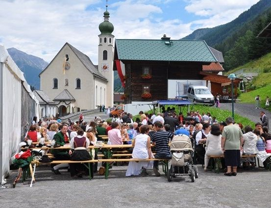 Eine große Gruppe von Menschen versammelt sich um Tische in einer Freiluftumgebung, wahrscheinlich für ein Festival. Im Hintergrund befinden sich Gebäude und Berge.