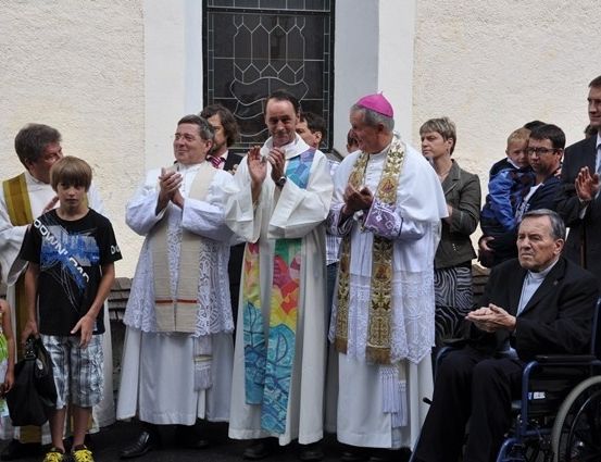Eine Gruppe von Menschen, darunter Priester und Laien, steht vor einer Kirche, klatscht und lächelt.