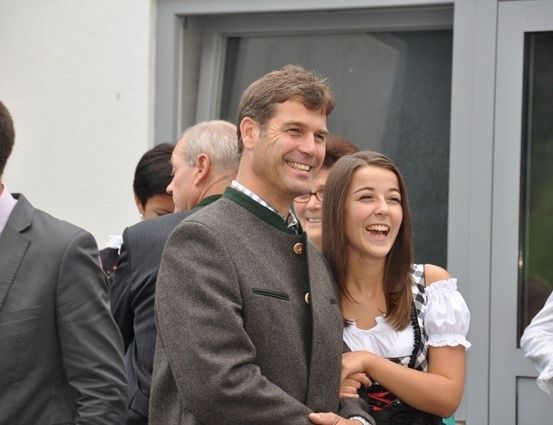 Ein Mann und eine Frau lächeln und stehen eng beieinander, möglicherweise bei einer gesellschaftlichen Veranstaltung. Der Mann trägt einen grauen Mantel mit grünem Kragen, und die Frau trägt ein weißes und schwarzes Kleid. Dahinter stehen mehrere Personen, von denen einige verschwommen sind.