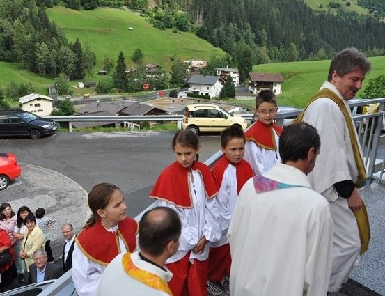 Ein Priester und zwei junge Messdiener sprechen mit einer Gruppe von Menschen, mit einem bergigen Hintergrund.