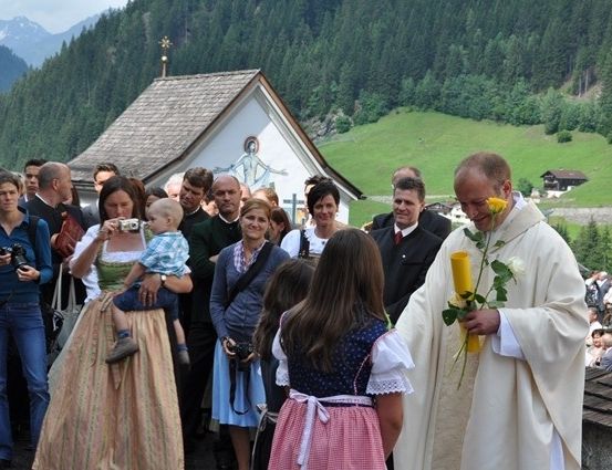 Ein Priester hält Blumen und eine Kerze, während sich eine Menschenmenge vor einer Kapelle in den Bergen versammelt.