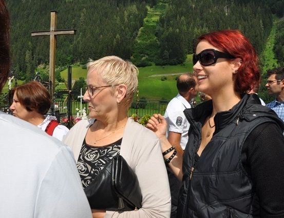 Eine Gruppe von Menschen steht draußen in der Nähe eines Holzkreuzes, wobei eine Frau in einer schwarzen Jacke lächelt und eine Frau mit Brille ernst aussieht.