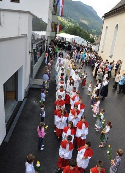 Eine große Gruppe von Menschen, darunter Kinder in roten und weißen Gewändern, geht in einer Prozession auf einer Straße. Sie halten Blumen und nehmen anscheinend an einer religiösen Veranstaltung teil.