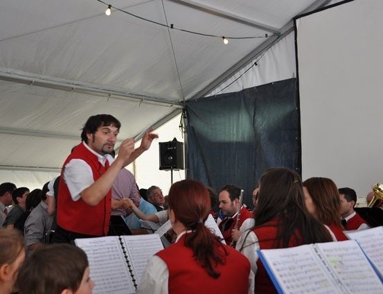 Ein Mann in einem roten Weste dirigiert eine Band in einem Zelt, während eine Menge zuschaut.