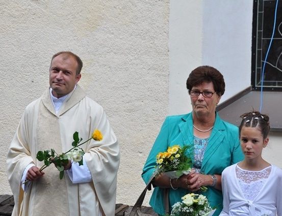 Ein Priester in einer weißen Robe hält einen Strauß weißer Rosen, neben einer Frau in einem blauen Blazer und einem jungen Mädchen in einem weißen Kleid, alle vor einem Gebäude.
