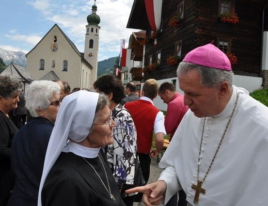 Ein Priester mit einem rosa Hut und eine Nonne in einem weißen Habit unterhalten sich vor einer Kirche mit einer grünen Kuppel und einem Turm.