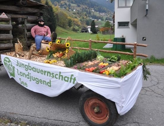 Ein Mann sitzt auf einem mit Gemüse und Sonnenblumen gefüllten Wagen, mit einem Holzzaun und einem Haus im Hintergrund.