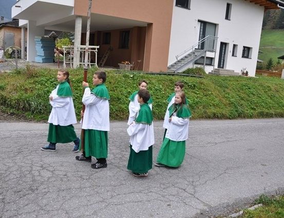 Eine Gruppe von Kindern in grünen und weißen Gewändern geht auf einer Straße vor einem Haus.