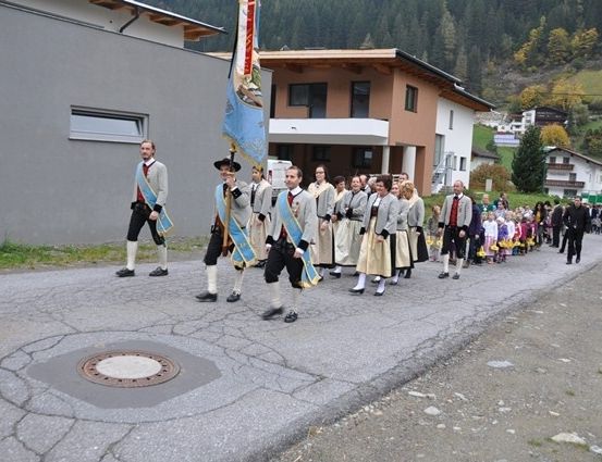 Eine Gruppe von Menschen in traditionellen Kostümen marschiert bei einer Parade auf einer Straße mit Gebäuden und Bergen im Hintergrund.