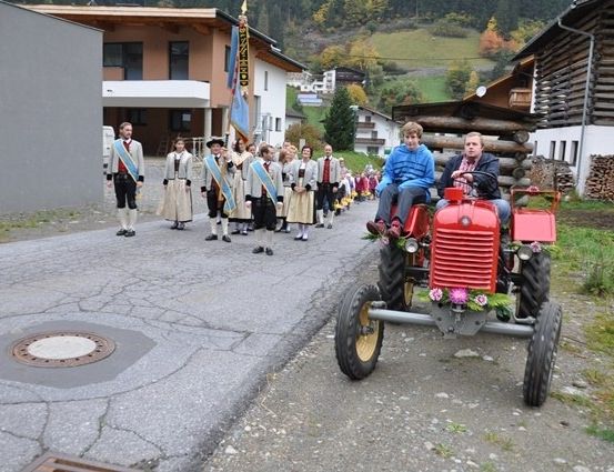 Eine Gruppe von Menschen in traditioneller Kleidung geht hinter einem roten Traktor mit Blumen darauf.