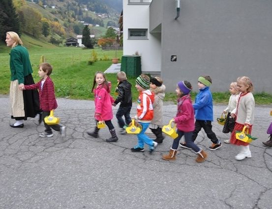 Eine Gruppe von Kindern, in Winterkleidung gekleidet, geht in einer Reihe und hält gelbe Körbe. Eine Frau in einem grünen Kleid führt sie an. Sie befinden sich auf einer Straße mit einem Gebäude und einem Grasfeld im Hintergrund.