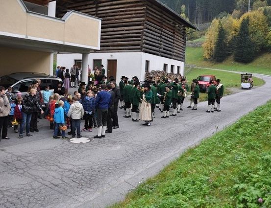 Eine Gruppe von Menschen in traditionellen grünen Uniformen steht in einer Reihe am Straßenrand, während andere vom Bürgersteig aus zusehen.