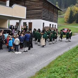 Eine Gruppe von Menschen in traditionellen grünen Uniformen steht in einer Reihe am Straßenrand, während andere vom Bürgersteig aus zusehen.