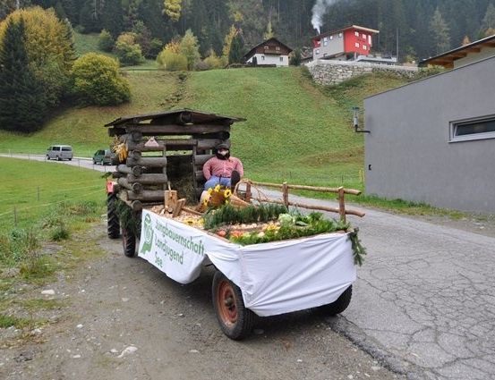 Eine Person mit Maske sitzt auf einem Traktoranhänger voller Gemüse und Blumen auf einer ländlichen Straße.