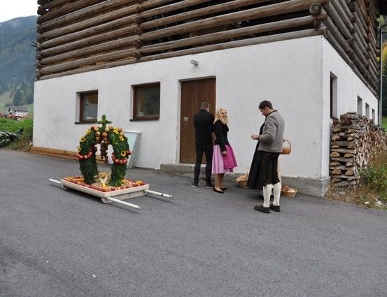 Drei Personen stehen vor einem rustikalen Haus mit Holzfassade und einer Blumenarrangement auf einem Karren davor.