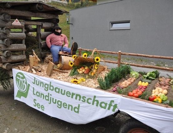 Ein hölzerner Wagen mit einem rustikalen Blockhüttendach zeigt eine Vielzahl von Gemüse und Früchten. Eine Person in einem roten karierten Hemd und Hut sitzt auf dem Wagen. Das Banner lautet 'Jungbauernschaft Landjugend See'.