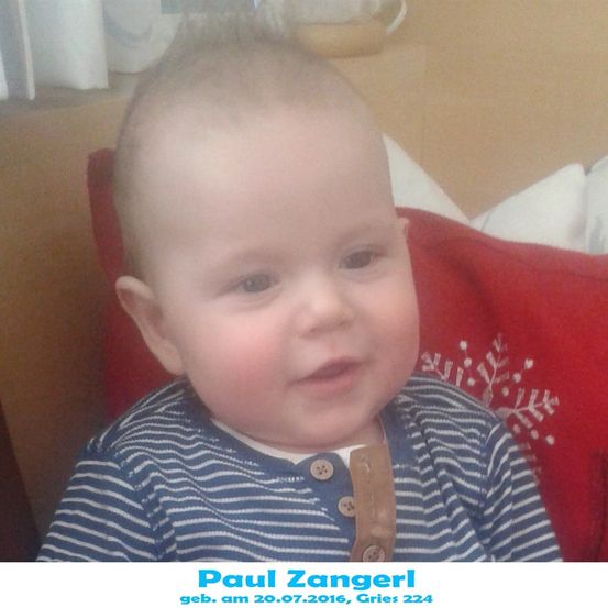 Ein Babyjunge mit kurzem Haar und einem blau-weiß gestreiften Shirt sitzt auf einem roten Kissen mit Schneeflocken-Design. Das Baby schaut mit einem leichten Lächeln nach vorne.