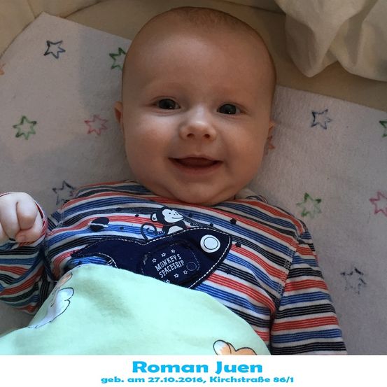 Ein lächelnder Babyjunge, der ein gestreiftes Shirt mit einem Affenaufnäher und einem blauen Raumschiffaufnäher auf der Brust trägt. Er liegt auf einem Bett mit einer weißen Decke und sternförmigen Dekorationen.