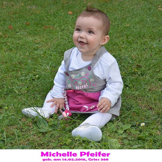 Ein Babymädchen sitzt auf dem Rasen, lächelt und schaut in die Kamera. Sie trägt ein graues Kleid mit einem rosa Rock und weißen Schuhen. Der Rasen ist grün und es gibt kleine weiße Blumen. Das Foto hat einen Wasserzeichen mit dem Namen Michelle Pfeifer.