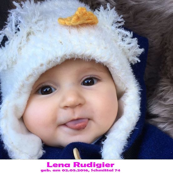 Ein Babymädchen namens Lena Rudigier, geboren am 2. Mai 2016, trägt einen weiß gefütterten Hut mit gelbem Schnabel und einem blauen Mantel. Sie streckt die Zunge heraus und schaut in die Kamera.