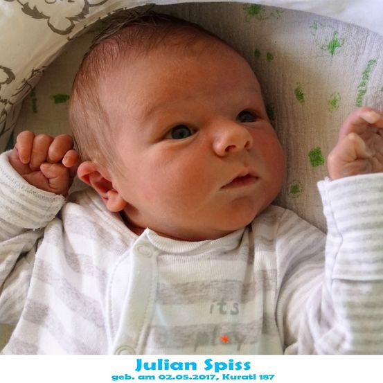 Ein neugeborener Junge namens Julian Spiss, geboren am 2. Mai 2017, liegt auf einem Bett mit den Händen in der Nähe seiner Ohren, trägt ein weißes gestreiftes Shirt mit dem Wort 'its' darauf.
