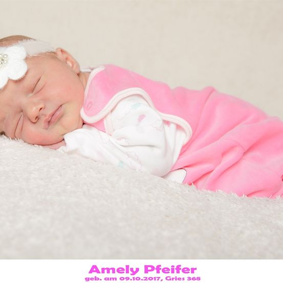 Ein Babymädchen schläft friedlich auf einem weißen Teppich. Sie trägt einen rosa Overall mit einem weißen Lätzchen und eine weiße Blumenhaarspange. Das Bild hat einen Wasserzeichen mit dem Text 'Amely Pfeifer' unten.