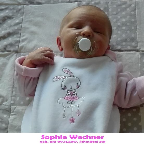 Ein neugeborenes Mädchen namens Sophie Wechner, geboren am 9. November 2017, schläft mit einem Schnuller im Mund. Sie trägt ein rosa Kaninchen-Lätzchen und liegt auf einer gepunkteten Decke.