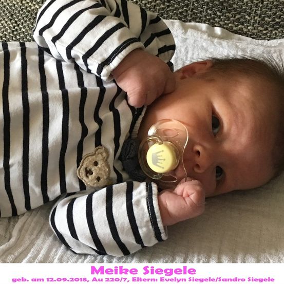 Ein Babyjunge mit einem Schnuller im Mund, auf einem weißen Tuch liegend, trägt ein schwarz-weiß gestreiftes Langarmshirt.