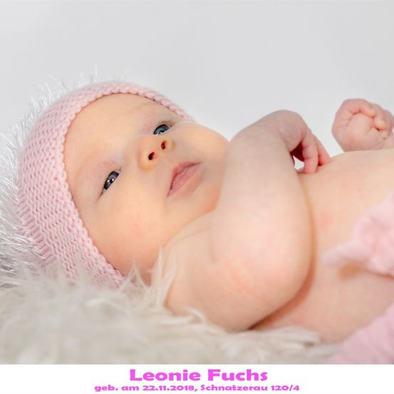 Ein Babymädchen mit blauen Augen trägt eine rosa gestrickte Mütze und liegt auf einer weißen Pelzdecke. Der Name des Babys, Leonie Fuchs, und ihr Geburtsdatum, 22.11.2018, sind unten angegeben.