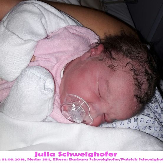 Ein neugeborenes Babymädchen schläft friedlich in einem Krankenhausbett, trägt ein rosa Outfit und einen klaren Schnuller. Sie hat lockiges Haar und ist in eine weiße Decke gewickelt. Das Bild ist vom 31. März 2018.
