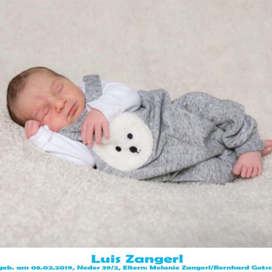 Ein neugeborener Babyjunge namens Luis Zangerl, geboren am 8. Februar 2019, schläft friedlich auf einem weißen Teppich. Er trägt ein graues Strampler mit einem weißen Bären-Plüschtier.
