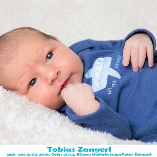 Ein Babyjunge namens Tobias Zangerl, geboren am 12. Februar 2019, liegt auf einer weißen Decke und trägt ein blaues Shirt mit einem Flugzeugdesign, während er seine Hand zum Mund hält.