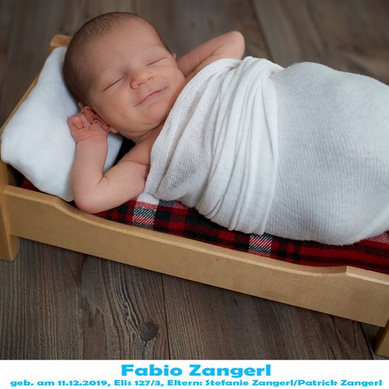 Ein Neugeborenes, in ein weißes Tuch gewickelt, schläft friedlich in einem Holzbettchen mit rot-schwarzem Karolaken. Das Baby liegt auf einem weißen Kissen und das Bettchen steht auf einem Holzboden.