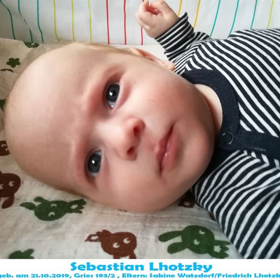 Ein Babyjunge namens Sebastian Lhotzky, geboren am 21. Oktober 2019, liegt auf einem Bett mit einer farbenfrohen Decke, die Schildkröten abbildet. Er trägt ein gestreiftes Shirt und scheint in die Kamera zu schauen.