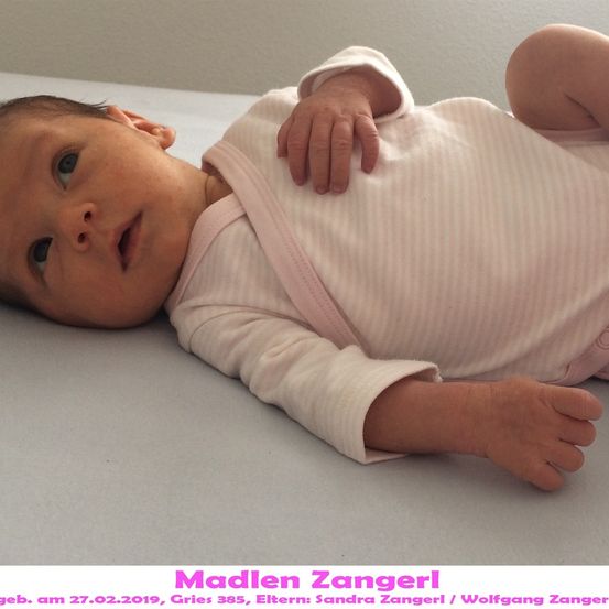 Ein Babymädchen mit blauen Augen, auf einem Bett liegend, trägt ein rosa Outfit. Das Bild hat einen Wasserzeichen mit dem Namen und Geburtsdatum des Babys.