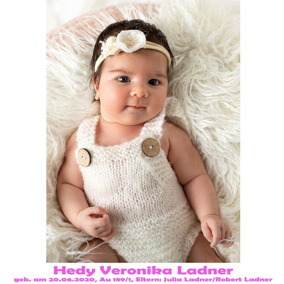 Ein Babymädchen liegt auf einer weißen Pelzdecke und trägt ein weißes gestricktes Overall mit zwei Holzknöpfen und einer weißen Blumenhaarspange. Das Baby lächelt und schaut in die Kamera. Das Bild hat einen Wasserzeichen mit dem Text 'Hed Veronika Ladner' unten.