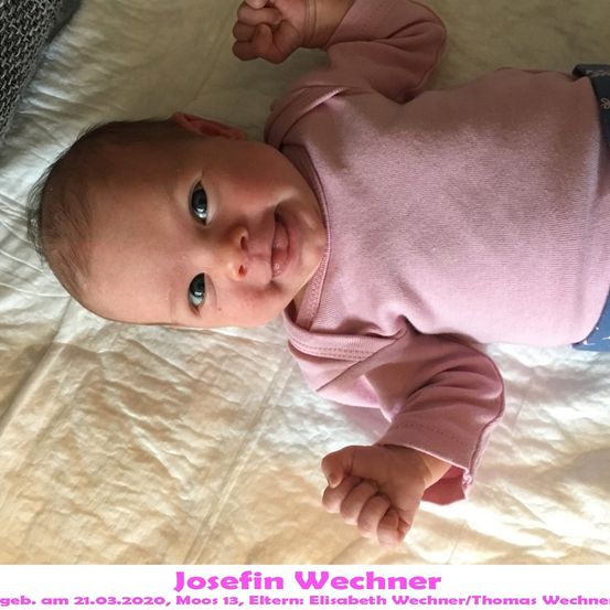 Ein Babymädchen namens Josefin Wechner, geboren am 21.03.2020, liegt auf einem Bett mit einem rosa Langarmshirt und blauer Hose. Sie hat blaue Augen und lächelt.