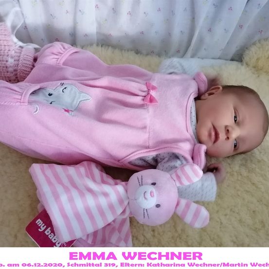 Ein Babymädchen liegt auf einer weichen Decke und trägt ein rosa Overall mit Katzendesign und hält ein rosa-weiß gestreiftes Plüschtier. Das Bild enthält einen Wasserzeichen mit dem Namen Emma Wechner.