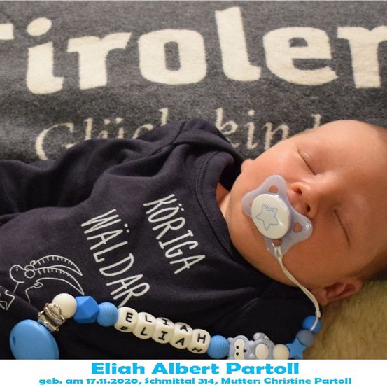 Ein Babyjunge namens Elijah schläft friedlich auf einer Decke mit einem Schnuller im Mund. Er trägt ein blaues Shirt mit dem Wort 'Waldar' und ein blau-weißes Armband mit seinem Namen.