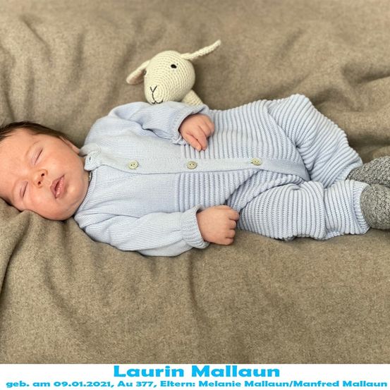 Ein Neugeborenes in blauer Kleidung liegt auf einer beigefarbenen Decke, mit einem gehäkelteten Schafsspielzeug in der Nähe. Das Baby schläft friedlich.