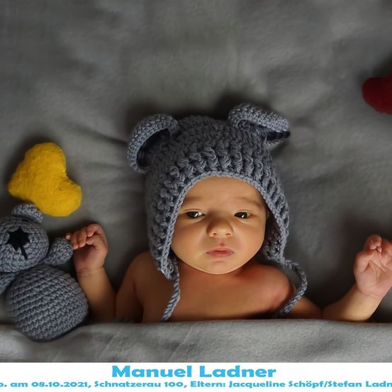 Ein Baby liegt auf einer grauen Oberfläche und trägt eine graue gehäkelte Bärenmütze, mit einem gelben Herz und einem grauen gehäkelten Bären in der Nähe. Der Name des Babys, Manuel Ladner, wird unten angezeigt.